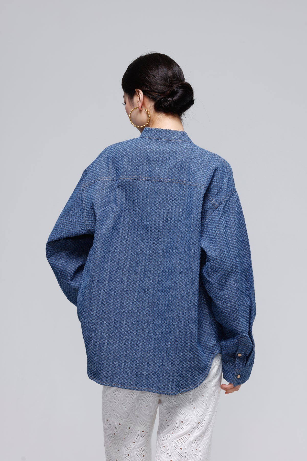 A woman shown from the back wearing a textured blue long-sleeve button-up shirt and white patterned pants, photographed against a solid light gray studio background.