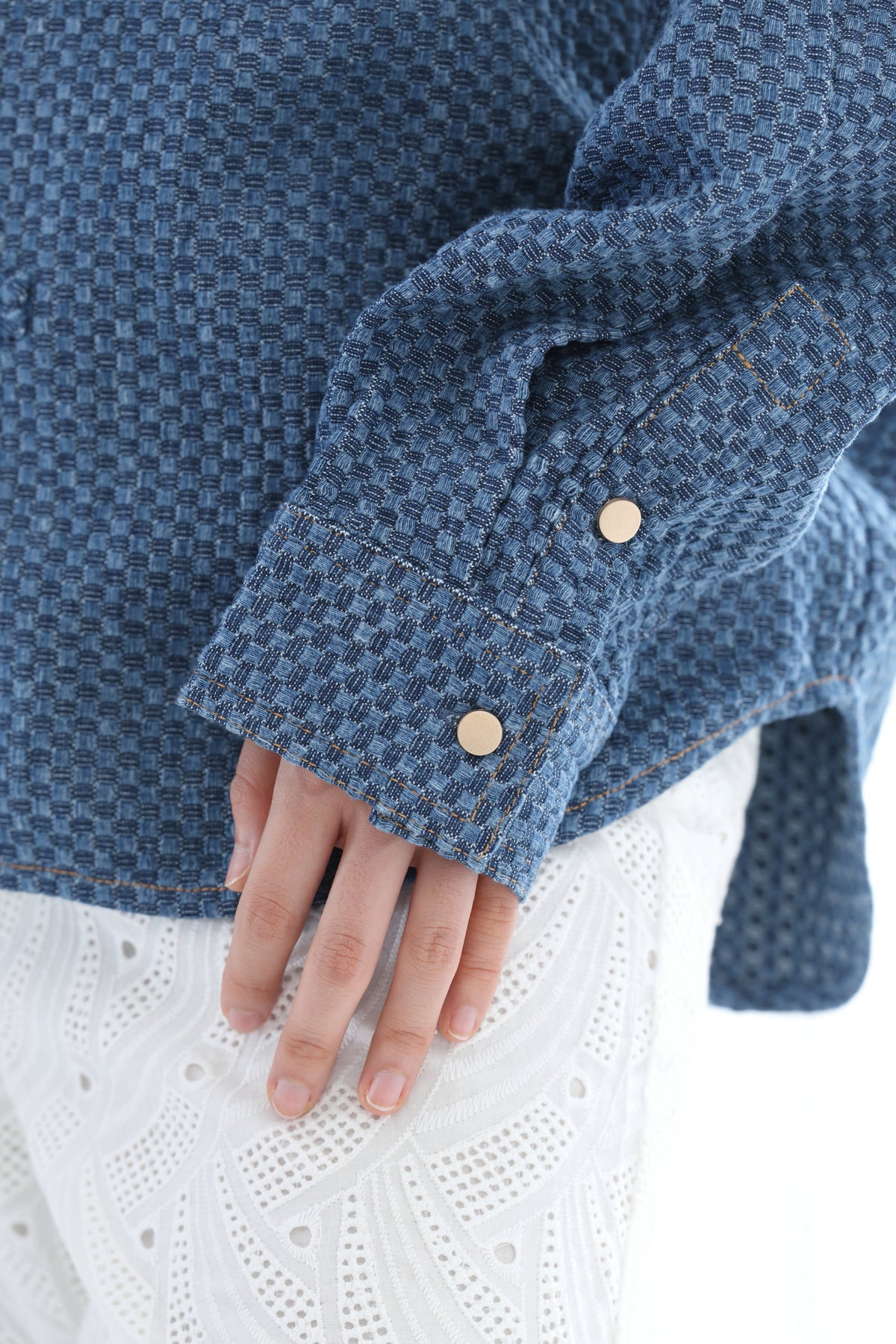 Close-up of a person wearing a textured blue woven jacket with gold-tone buttons and a white eyelet knit top underneath, against a clean white background.