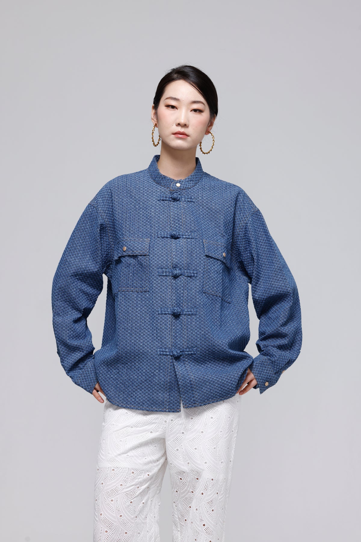 A woman modeling a blue textured long-sleeve jacket with traditional Chinese-style buttons and flap pockets, paired with white pants, against a solid gray studio background.