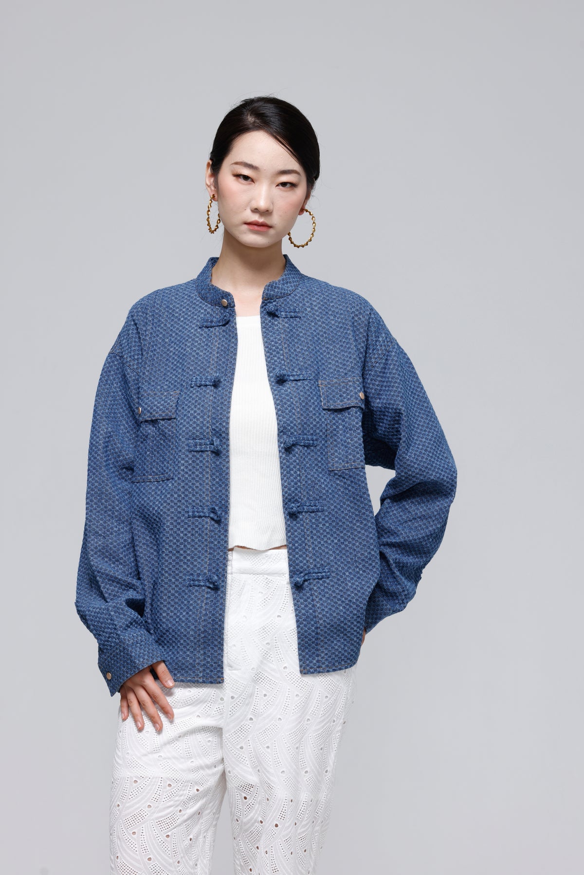 A woman modeling a blue textured jacket with traditional Chinese frog closures, worn over a white top and white pants, against a solid gray studio background.