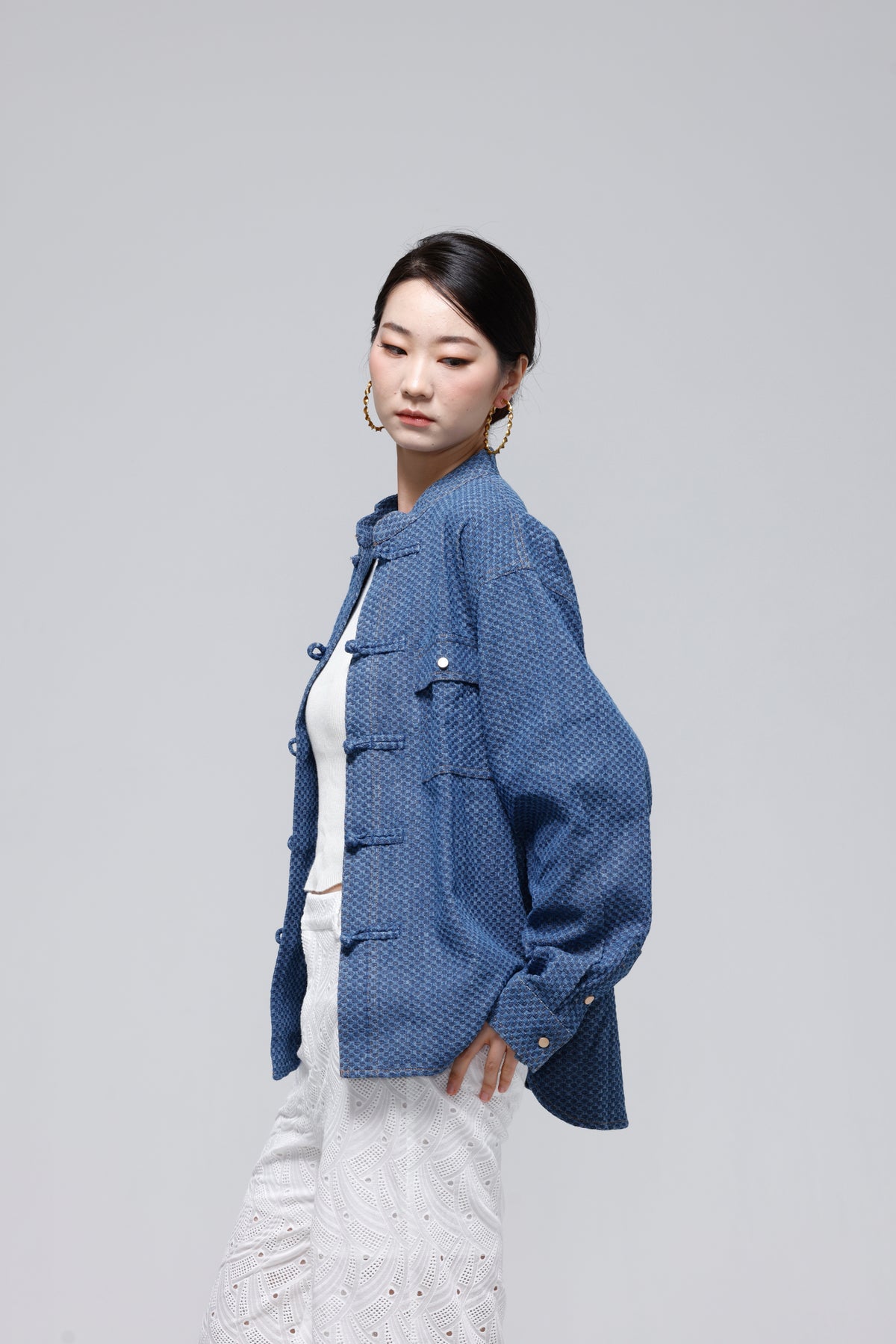 A woman modeling a blue textured jacket with traditional Chinese-style frog closures, worn over a white top and white patterned pants, set against a solid light gray background.