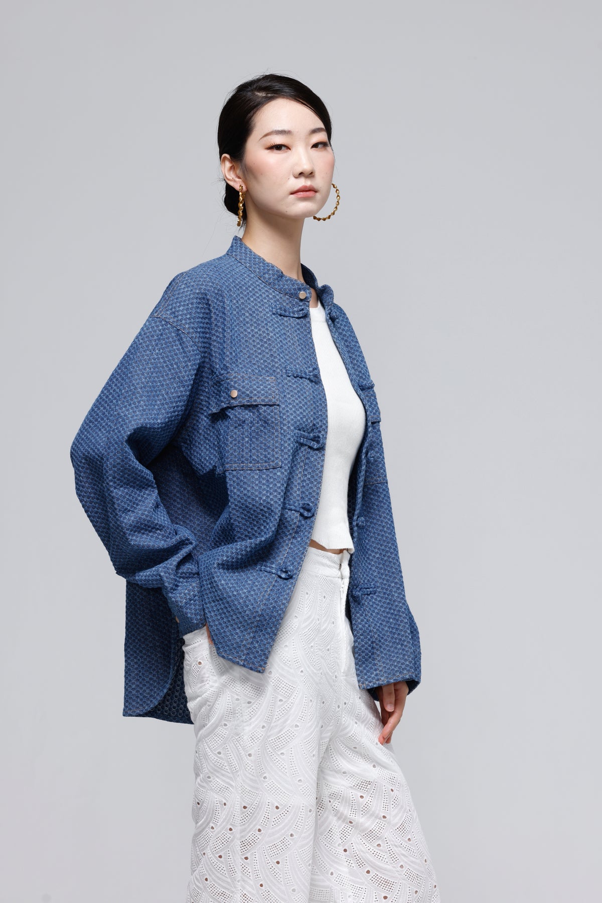 A woman modeling a textured blue jacket with mandarin collar and button closures, styled over a white top and white patterned trousers, against a solid gray studio background.