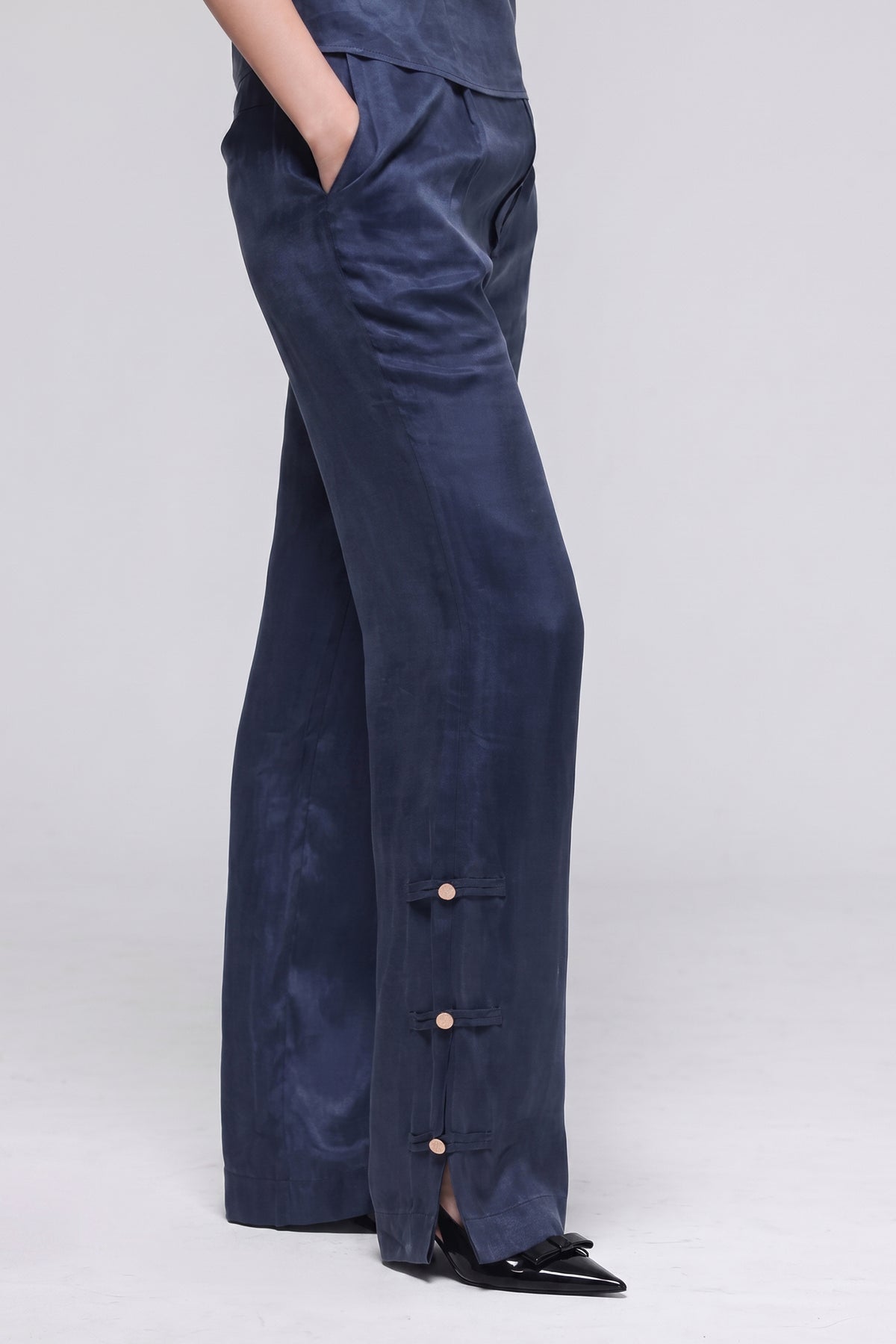 A person wearing navy wide-leg trousers with a high waist, side pockets, and three decorative gold buttons near the hem, paired with black pointed flats against a light gray studio background.