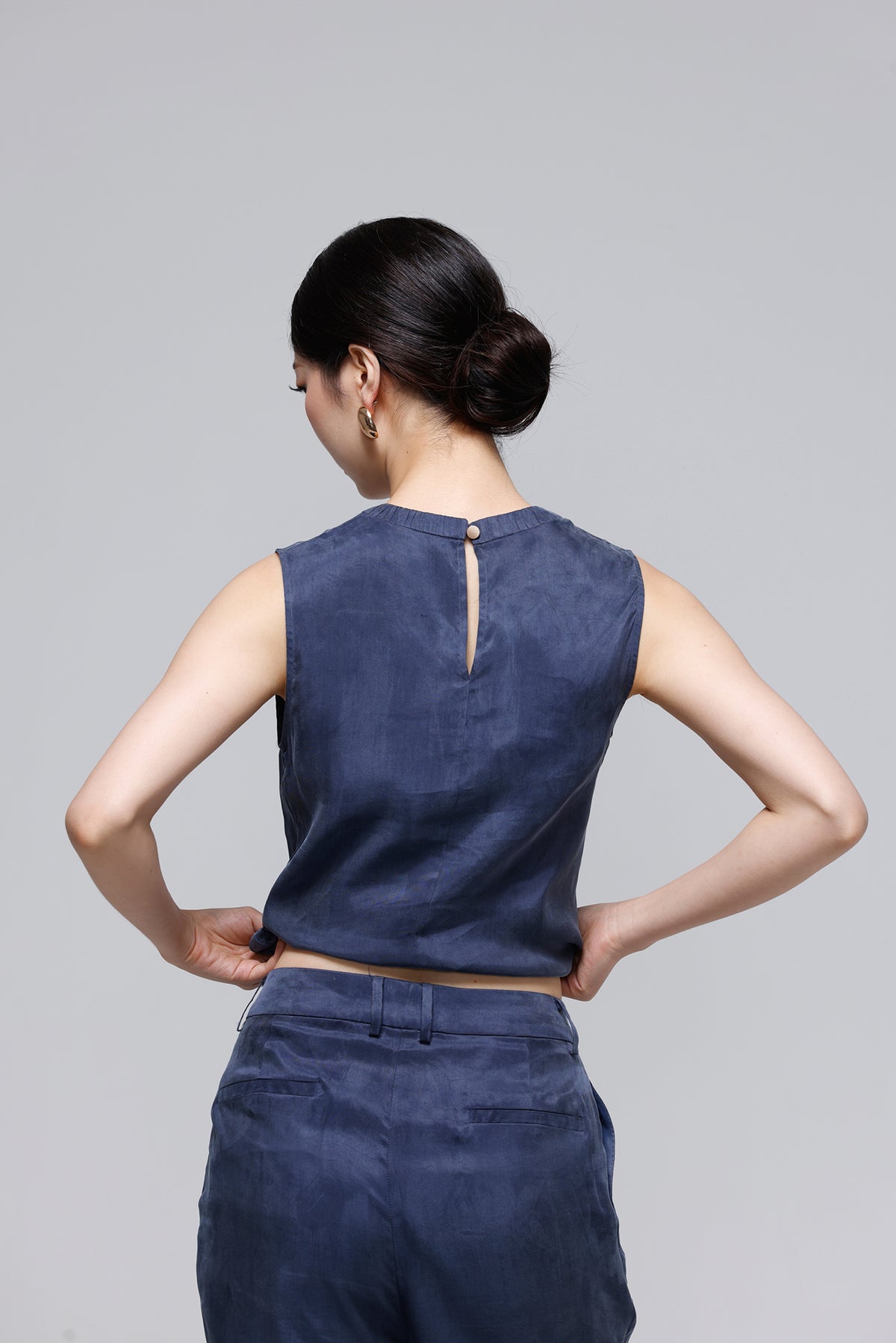 A woman shown from the back wearing a sleeveless navy blue top with a small back slit and matching navy pants, photographed against a plain light gray studio background.