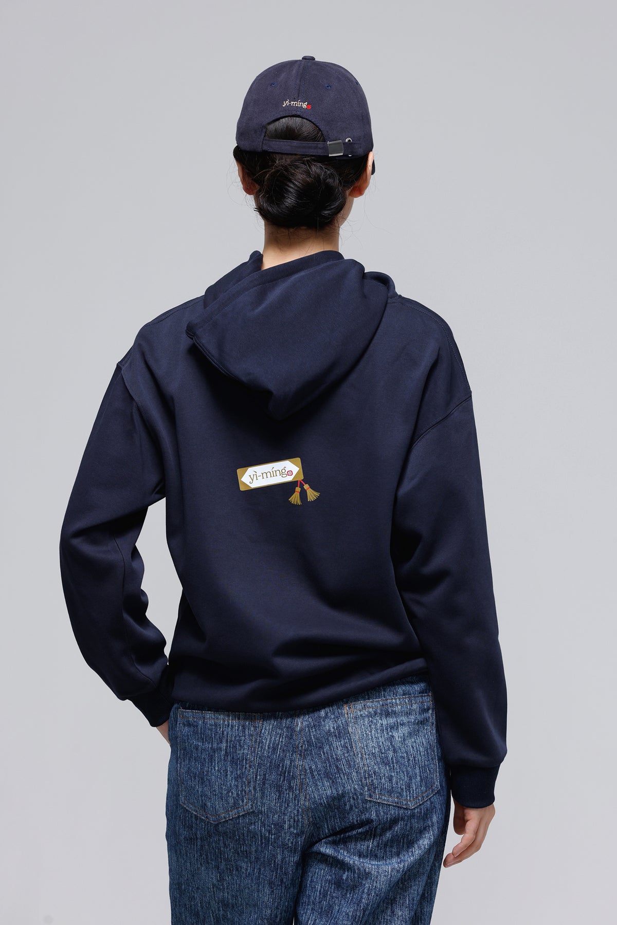 A person shown from the back wearing a navy blue hoodie with a small yellow and white graphic patch on the back, paired with blue jeans and a navy baseball cap, against a light gray studio background.