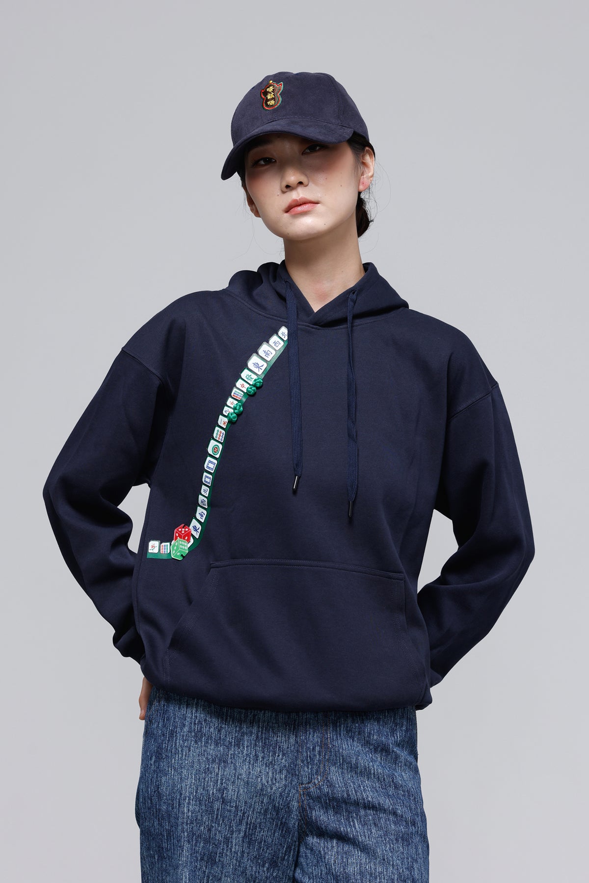 A model wearing a navy blue hoodie with a colorful diagonal graphic print on the front, paired with a matching navy blue baseball cap and blue jeans, against a solid gray studio background.