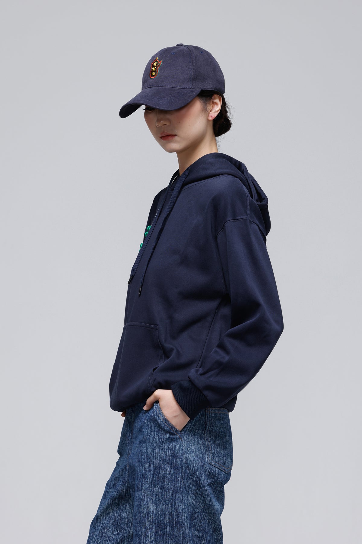 A model wearing a navy blue hoodie, a matching navy baseball cap with a small embroidered logo, and blue denim jeans, shown from a side profile against a solid gray studio background.