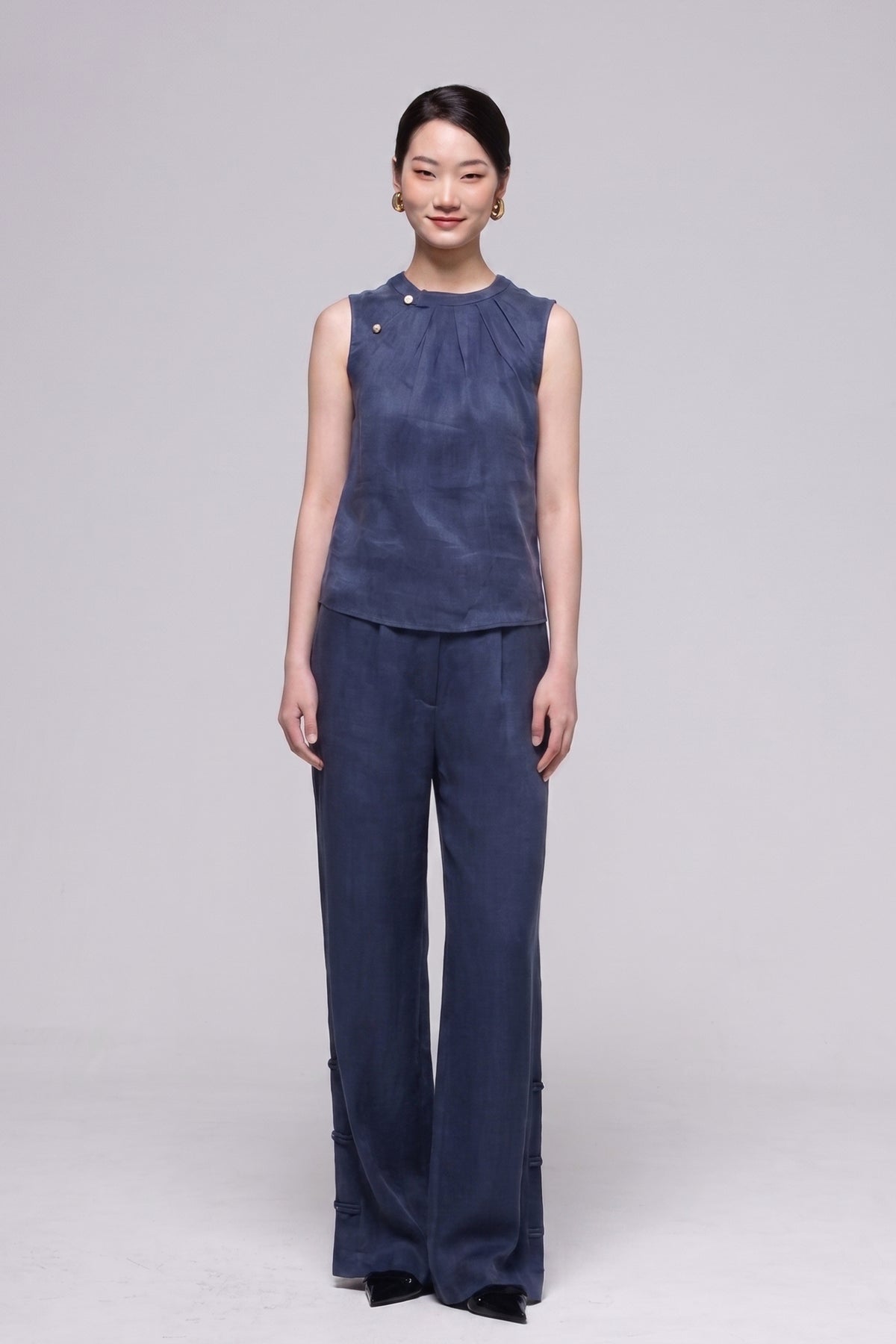 A woman modeling a matching navy blue two-piece outfit consisting of a sleeveless high-neck top with decorative buttons and matching wide-leg trousers, shot against a neutral light gray studio background.