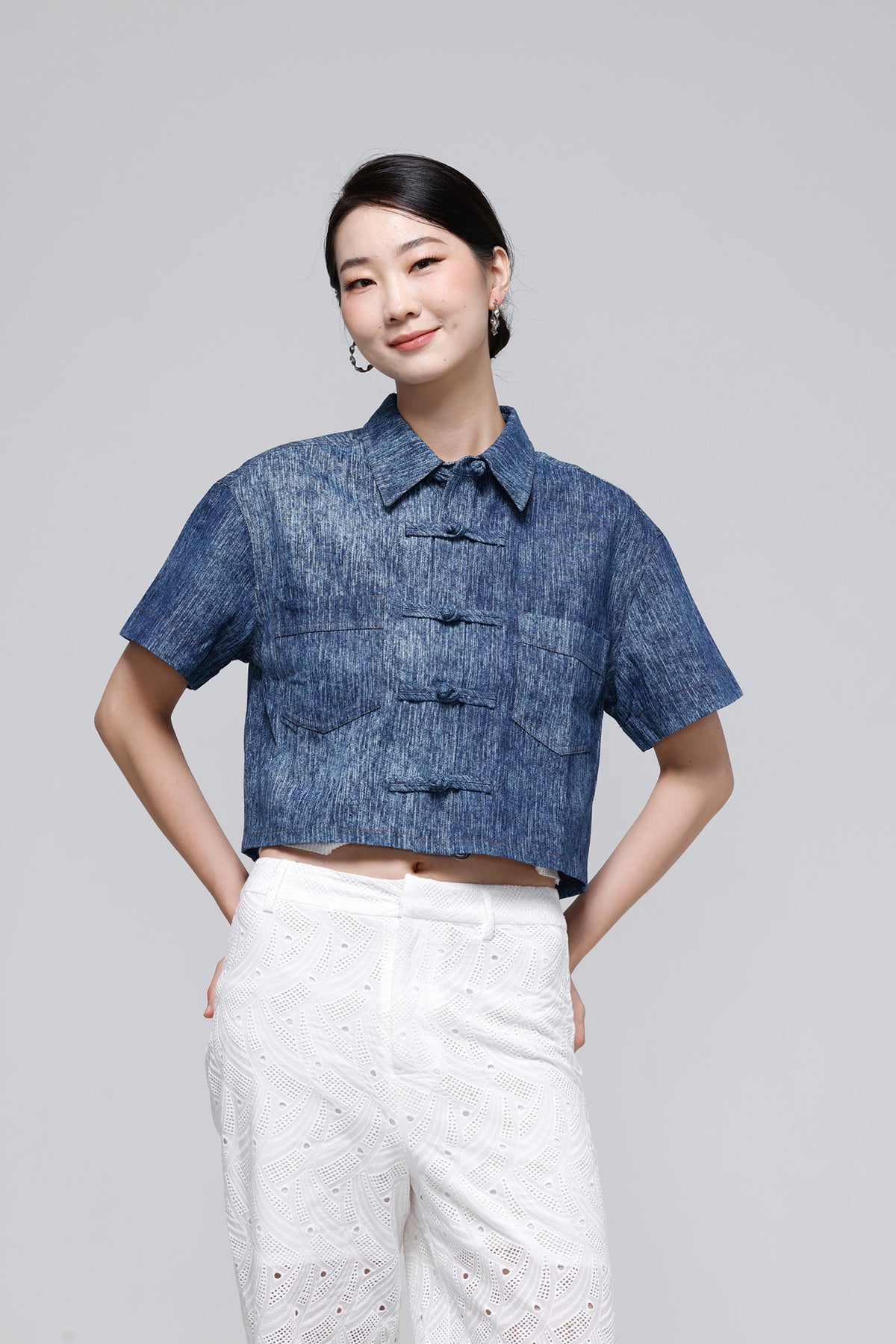 A woman modeling a short-sleeve cropped button-front shirt in a textured blue fabric, paired with white textured pants, against a solid light gray background.