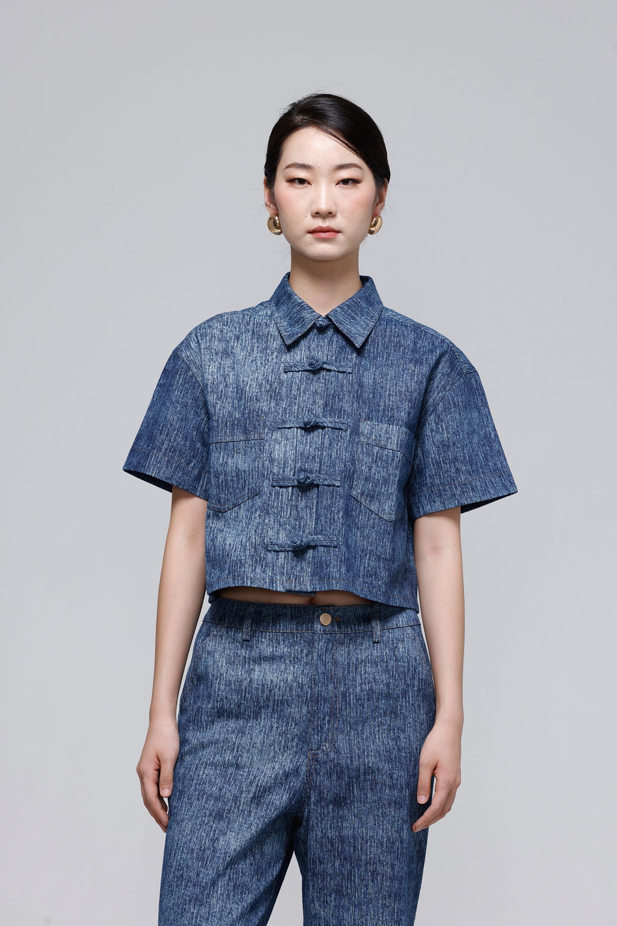 A model wearing a cropped short-sleeve button-front top and matching trousers in a blue denim-like fabric, photographed against a plain light gray studio background.