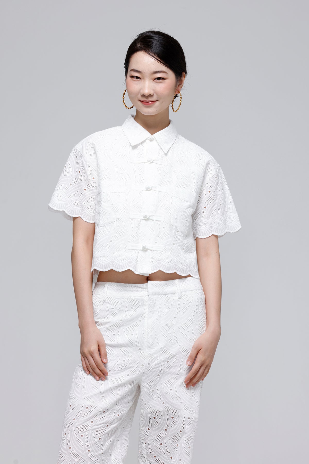 A woman modeling a white short-sleeve cropped blouse and matching white trousers with eyelet embroidery, photographed against a plain light gray studio background.