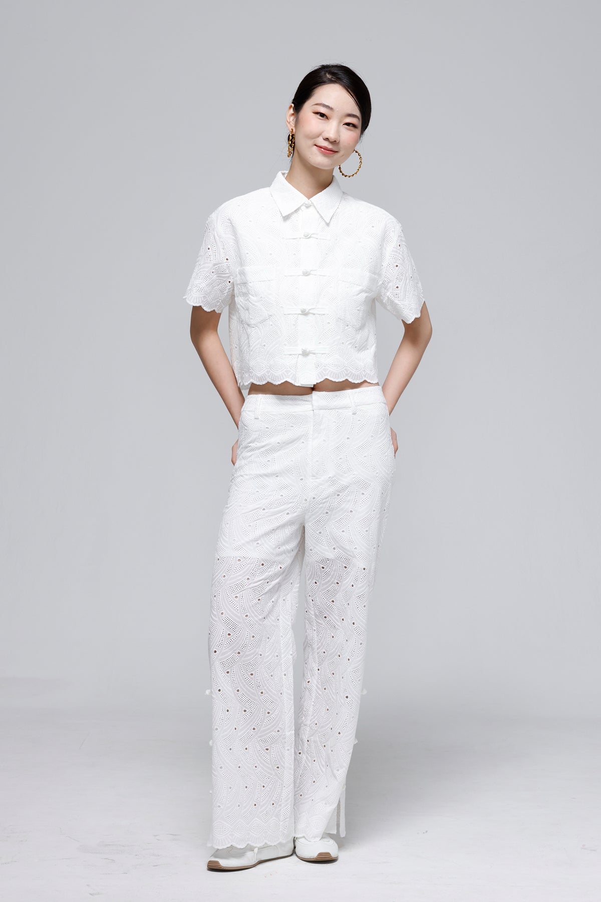 A woman modeling a white short-sleeve cropped button-up shirt and matching white wide-leg trousers with eyelet embroidery, photographed against a light gray studio background.