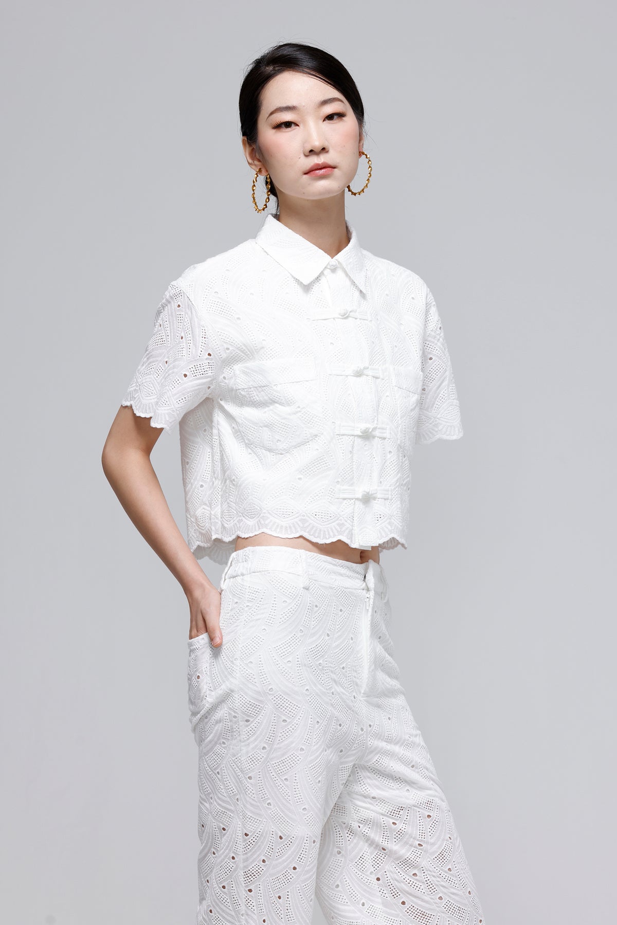 A woman modeling a white short-sleeve cropped blouse with scalloped edges and matching white wide-leg trousers, photographed against a light gray studio background.