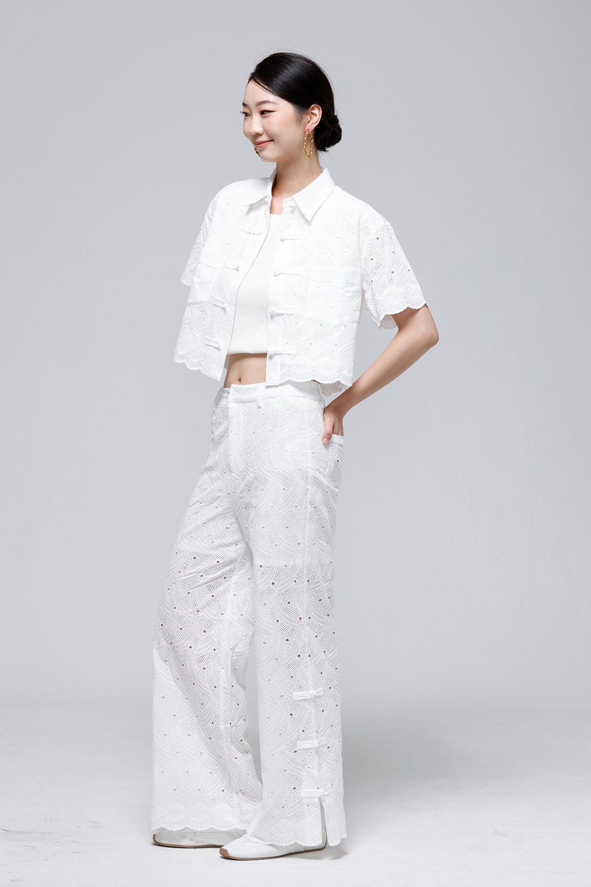 A woman modeling a white eyelet two-piece outfit consisting of a short-sleeve cropped button-front jacket and matching wide-leg pants, photographed against a light gray studio background.