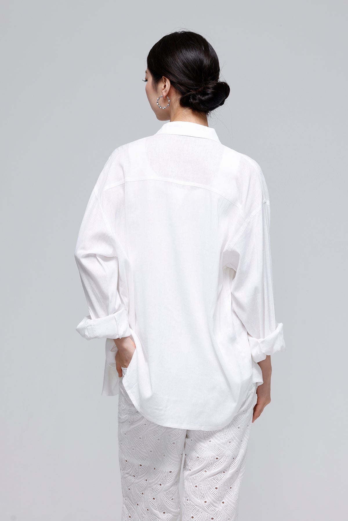 A woman shown from the back wearing an oversized white long-sleeve button-up shirt with rolled cuffs and white eyelet pants, photographed against a light gray studio background.
