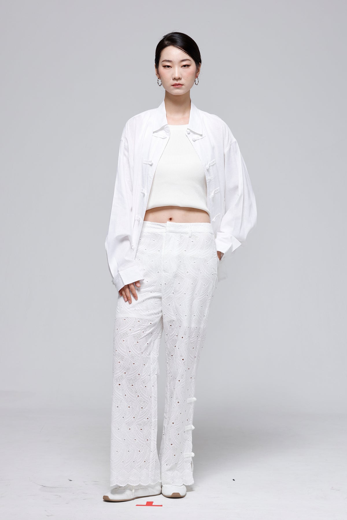 A model stands against a light gray studio background wearing a full white outfit consisting of a white cropped tank top, an open white button-up shirt, white wide-leg trousers with eyelet embroidery, and white sneakers.
