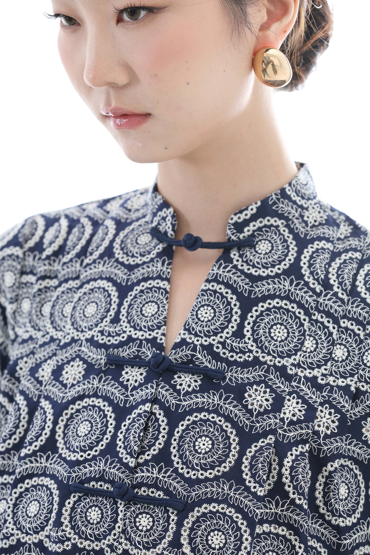 Close-up of a woman wearing a navy blue and white embroidered top with a mandarin collar and button closure, paired with a gold earring against a white background.