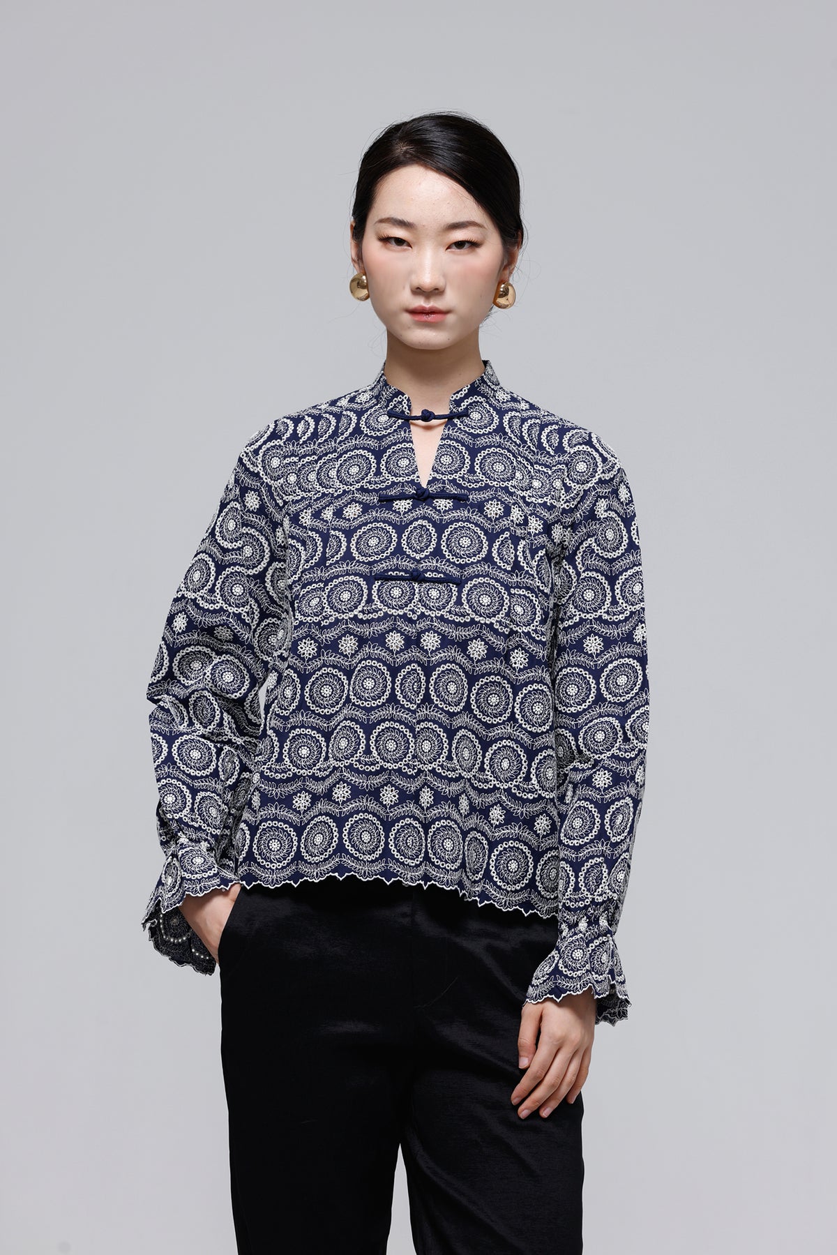 A woman modeling a navy blue long-sleeve blouse with a white circular mandala pattern and a keyhole cutout at the collar, paired with black pants, against a solid gray studio background.