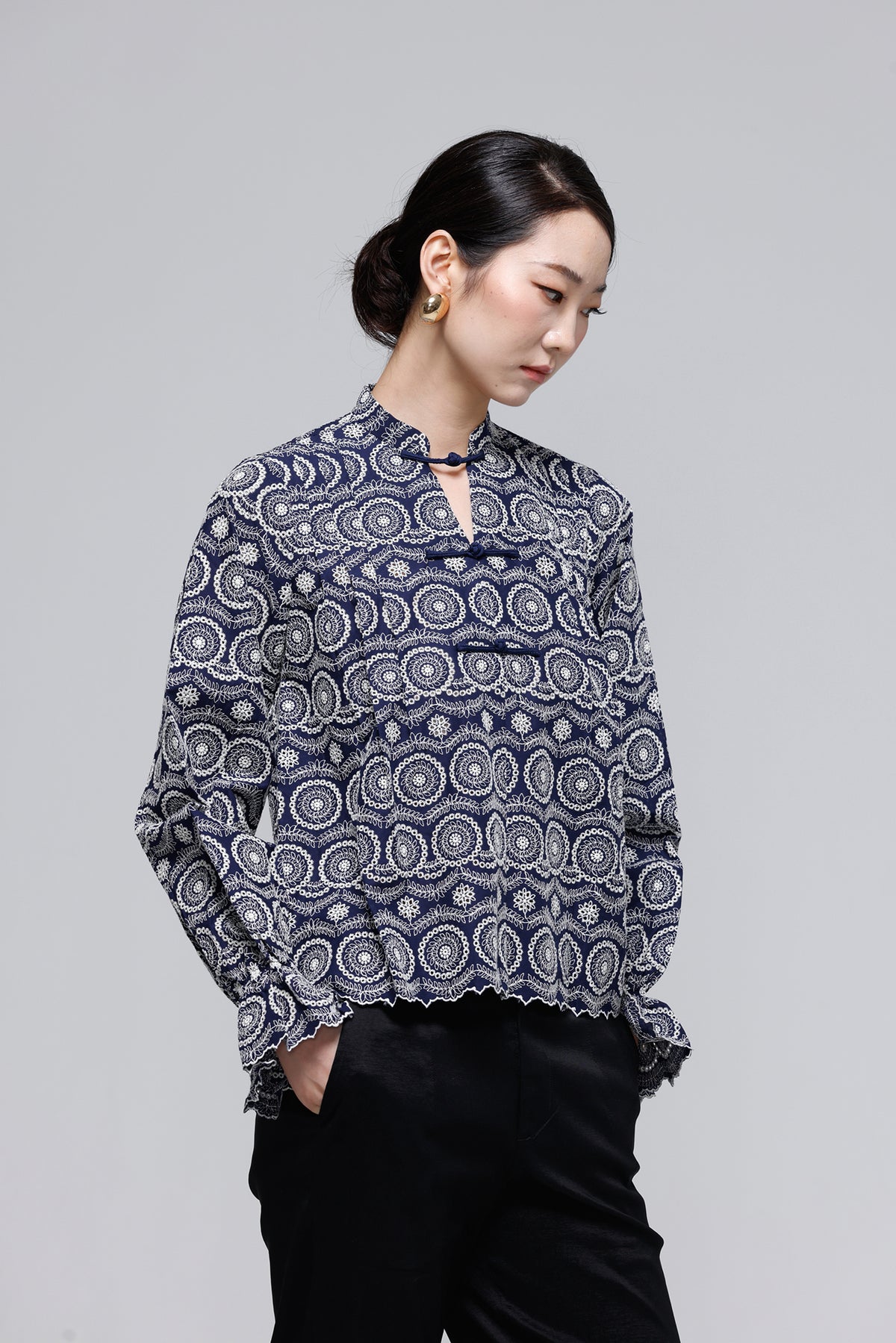 A woman wearing a navy blue long-sleeve blouse with a white circular mandala pattern and black pants, standing against a solid gray studio background.