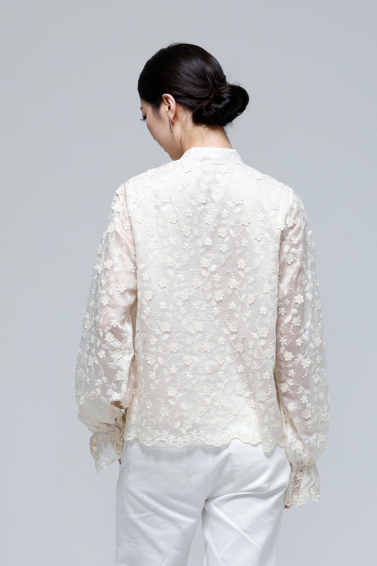 A woman viewed from the back wearing a cream-colored floral lace blouse with sheer long sleeves and white pants, set against a solid light gray studio background.