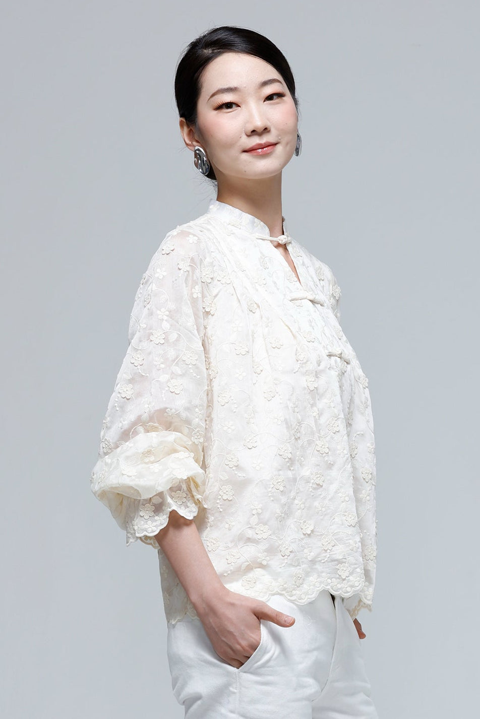A woman modeling a cream-colored, textured lace blouse with three-quarter ruffled sleeves and white pants, standing against a solid light gray studio background.