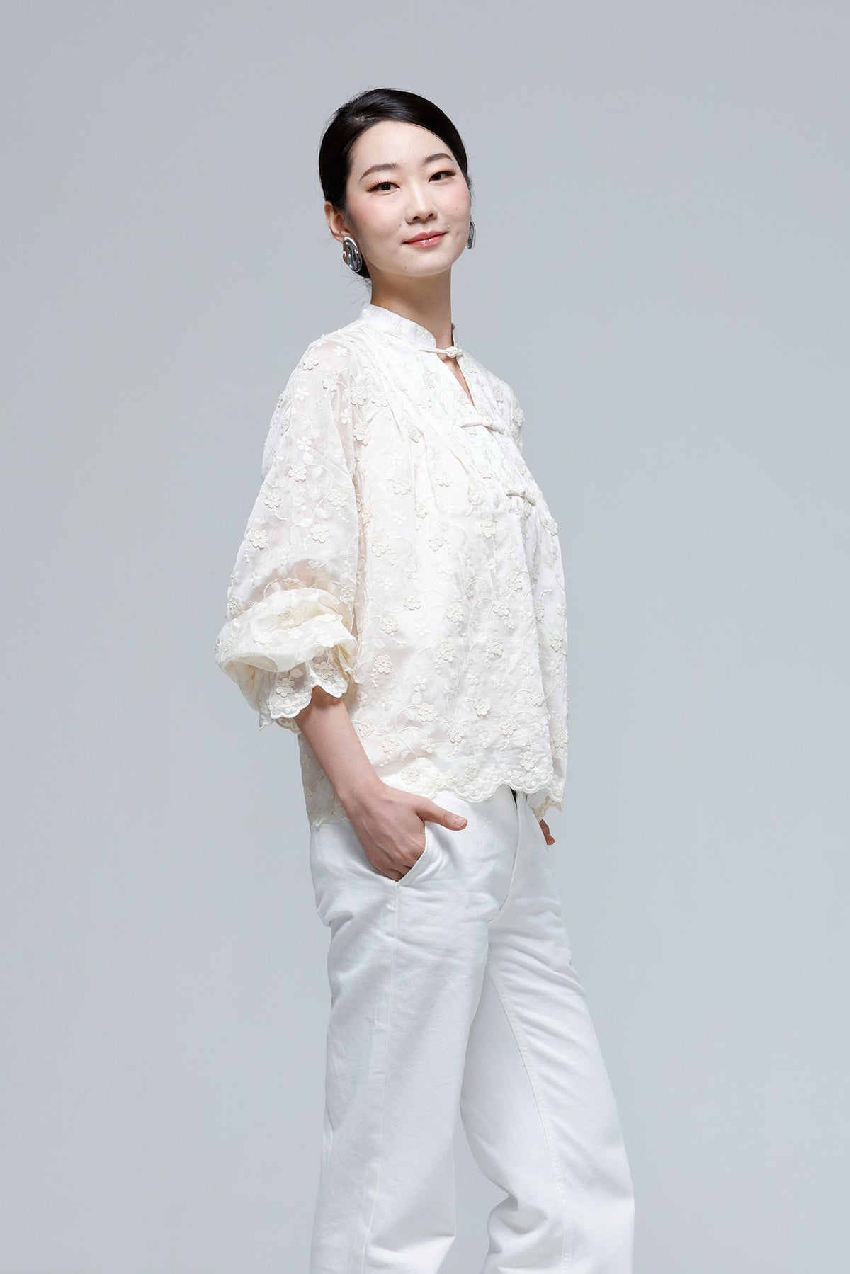 A woman modeling a cream-colored, textured lace blouse with three-quarter ruffled sleeves and white pants, standing against a solid light gray studio background.