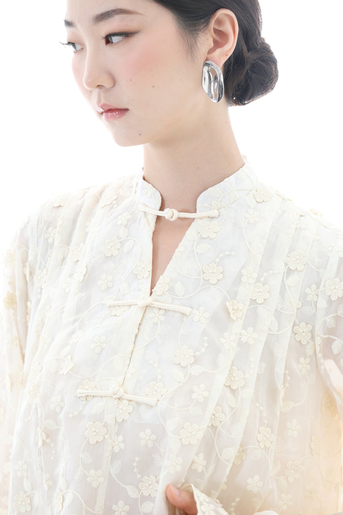 A close-up of a woman wearing a cream-colored, semi-sheer embroidered blouse with floral patterns and traditional Chinese-style frog closures, paired with a matching cream necklace and a silver hoop earring.