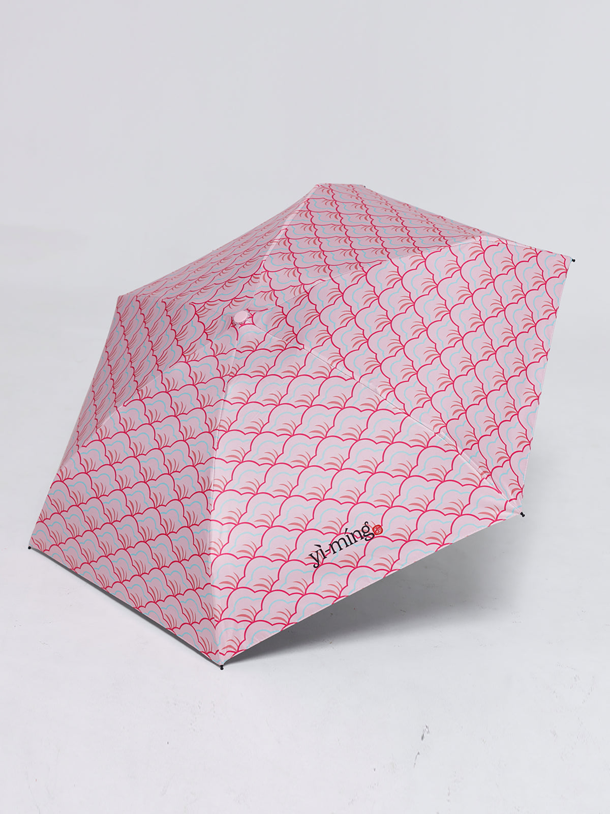 A compact folding umbrella with a pink repeating wave pattern, shown open on a light gray studio background. The brand name 'Yiming' is printed near the lower right section of the canopy.