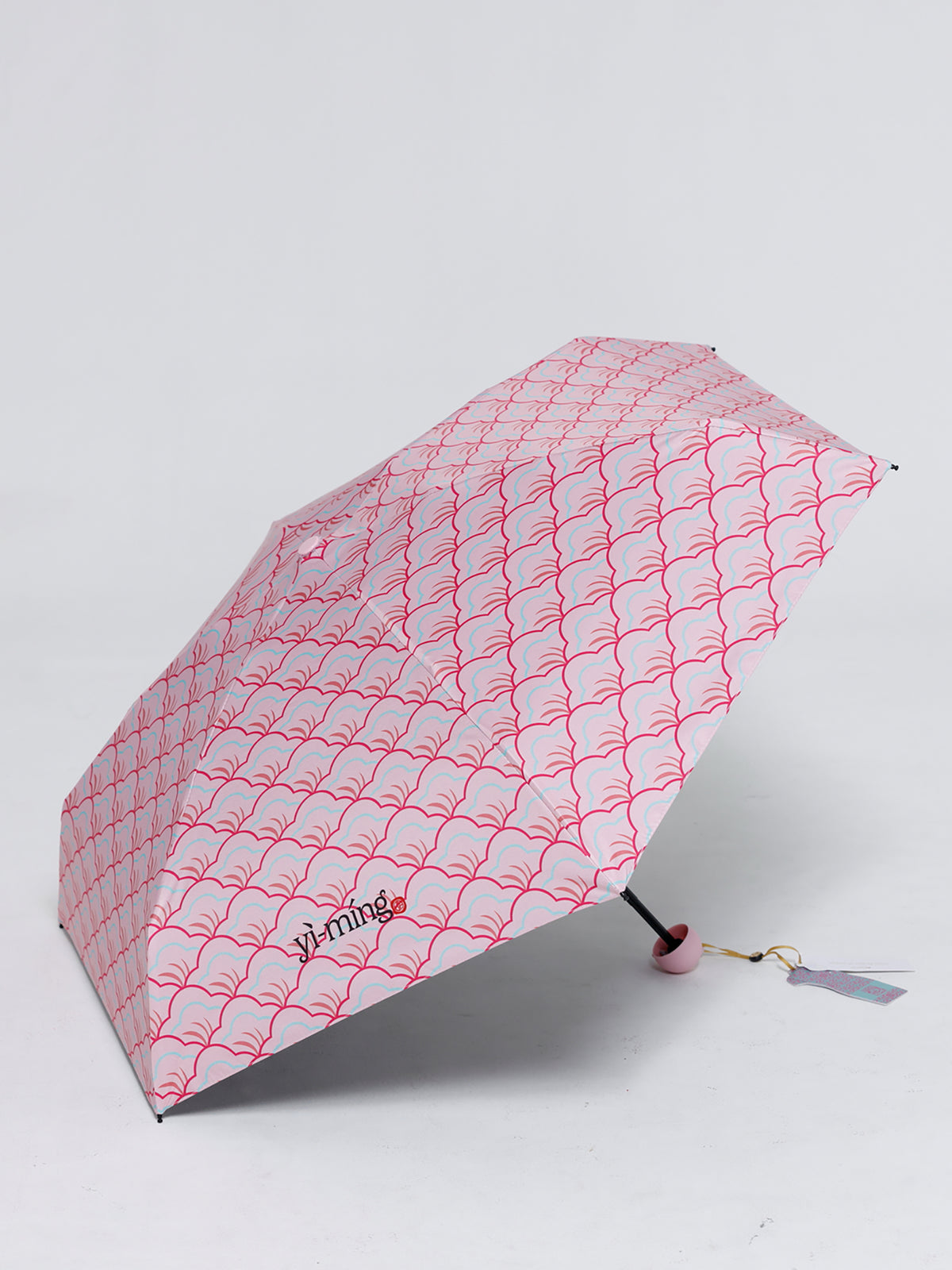A compact folding umbrella with a white canopy patterned with a pink wavy geometric design. The brand name 'yi ming' is printed in black near the lower left of the canopy.