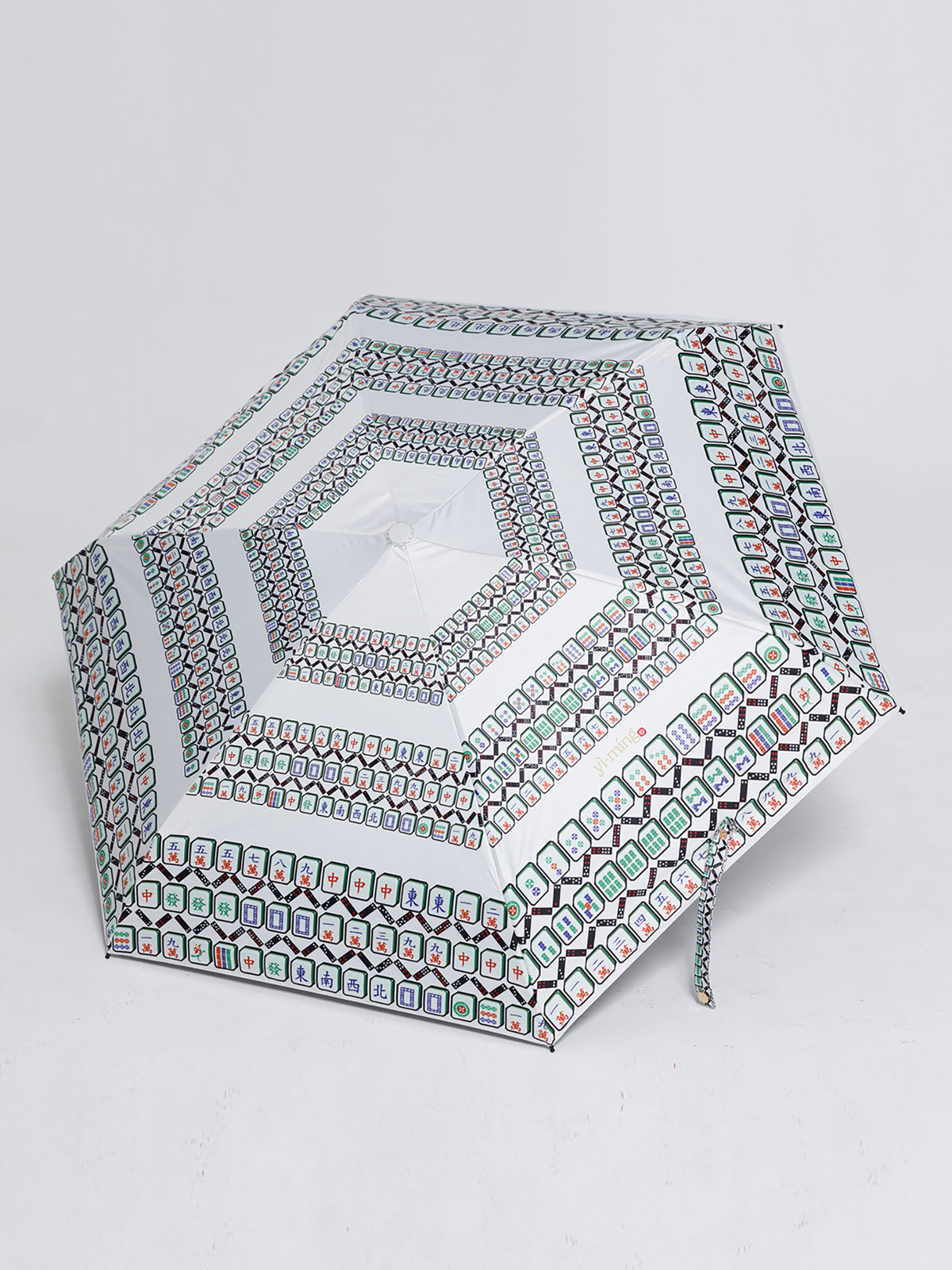 A compact, hexagonal umbrella with a white base and a dense, colorful geometric pattern of green, red, and blue motifs, shown on a light gray background.