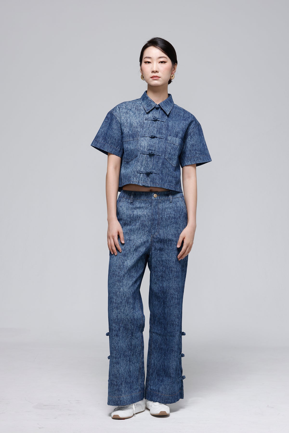 A model stands against a light gray studio background wearing a matching blue denim-like two-piece outfit consisting of a short-sleeve cropped button-front top and wide-leg pants with decorative side buttons.