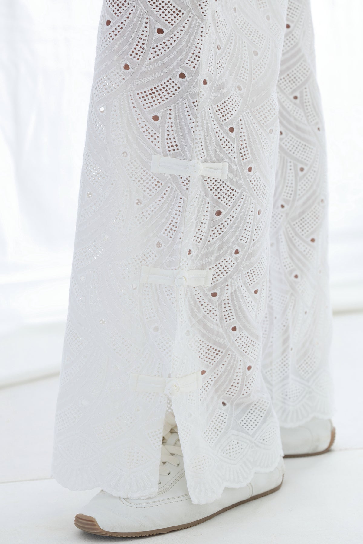White eyelet lace wide-leg pants with decorative bow details, shown on a model wearing white sneakers.