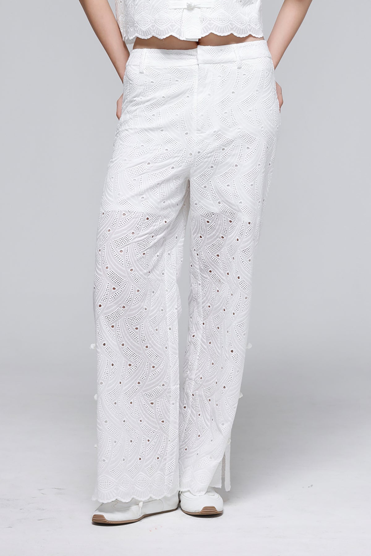 White wide-leg trousers with intricate eyelet embroidery and scalloped hems, shown on a model against a light gray studio background.