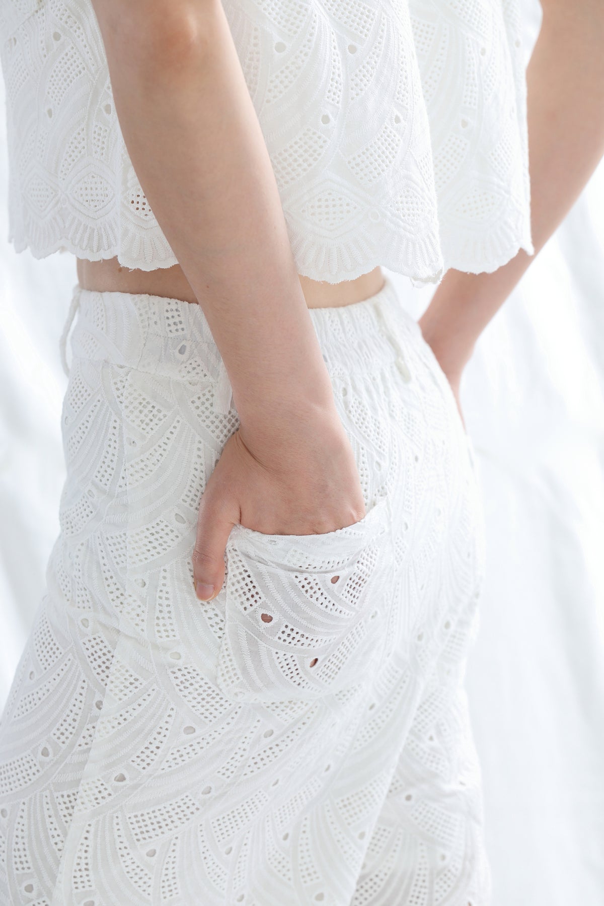 A close-up of a person wearing a white eyelet lace top and matching white eyelet pants, with one hand in a pocket. The background is a soft white fabric.