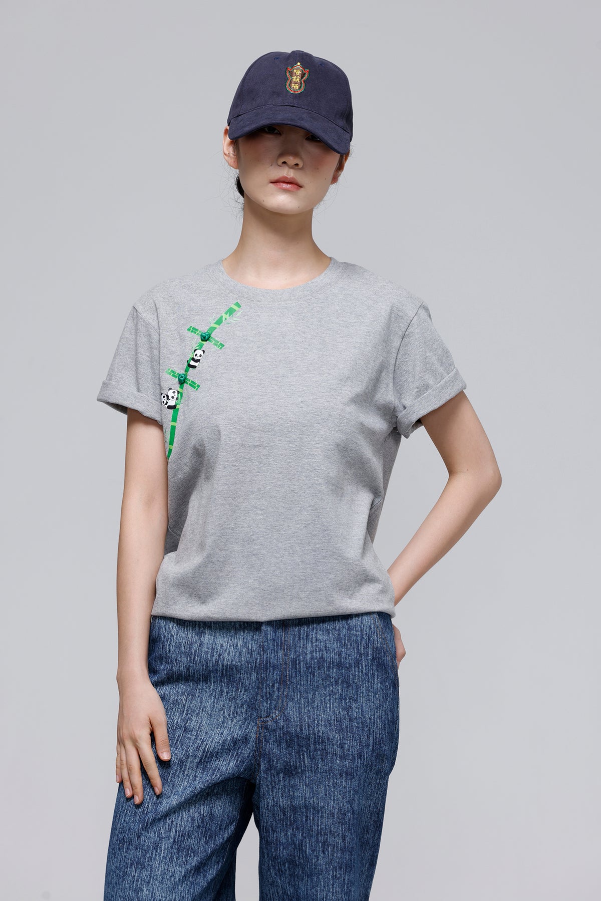 A model wearing a light gray crewneck t-shirt with a green bamboo graphic on the left chest, paired with blue jeans and a navy blue baseball cap, against a solid gray studio background.