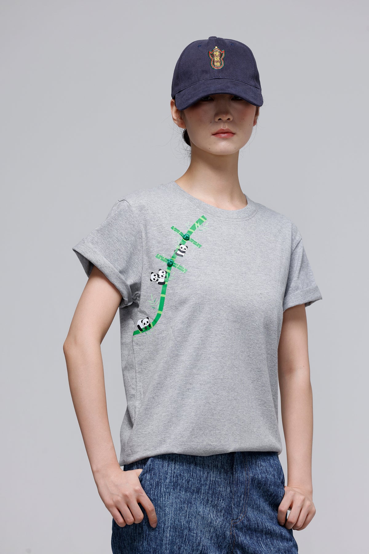A model wearing a light gray crewneck t-shirt with a green bamboo and panda graphic on the front, paired with a navy blue baseball cap and blue jeans, against a solid gray studio background.