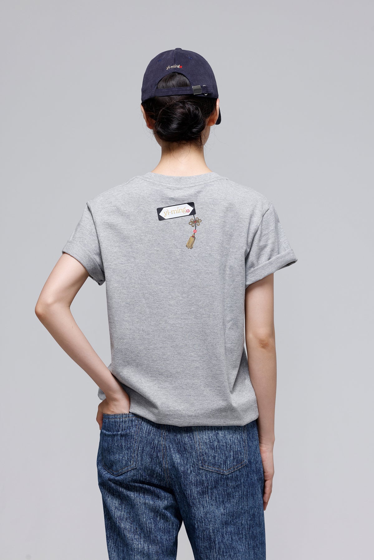 A person shown from the back wearing a light gray short-sleeve t-shirt with a small graphic on the upper back, blue jeans, and a dark navy baseball cap, against a solid gray studio background.