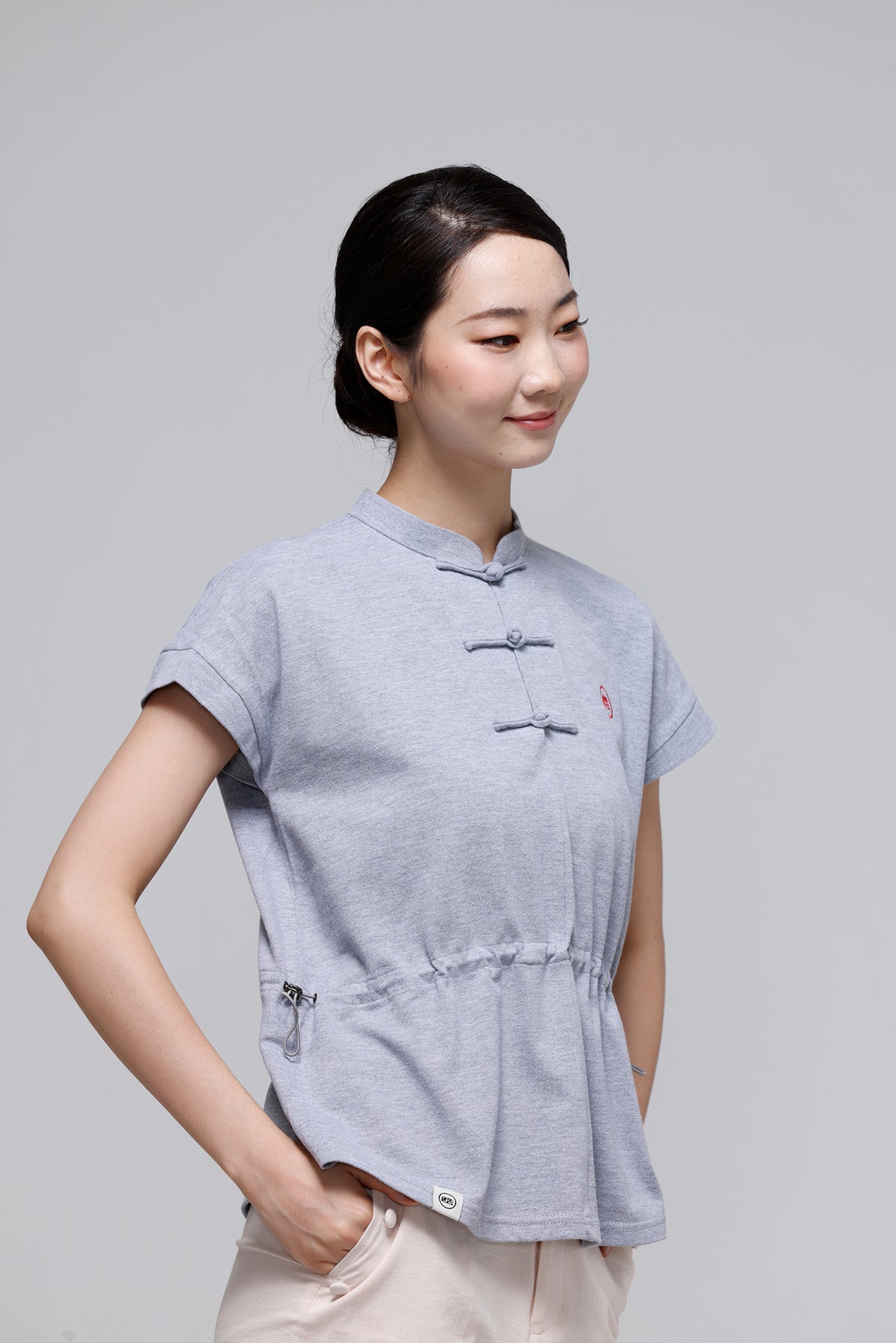 A woman wearing a light gray short-sleeve top with traditional Chinese-style frog closures and a drawstring waist, paired with beige pants, against a solid gray studio background.
