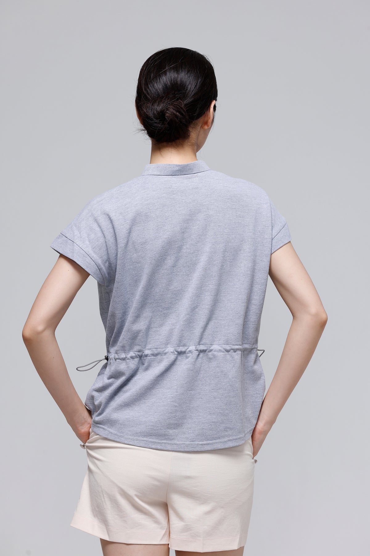 A woman shown from the back wearing a light gray short-sleeve top with a drawstring waist and light beige shorts, against a plain light gray studio background.