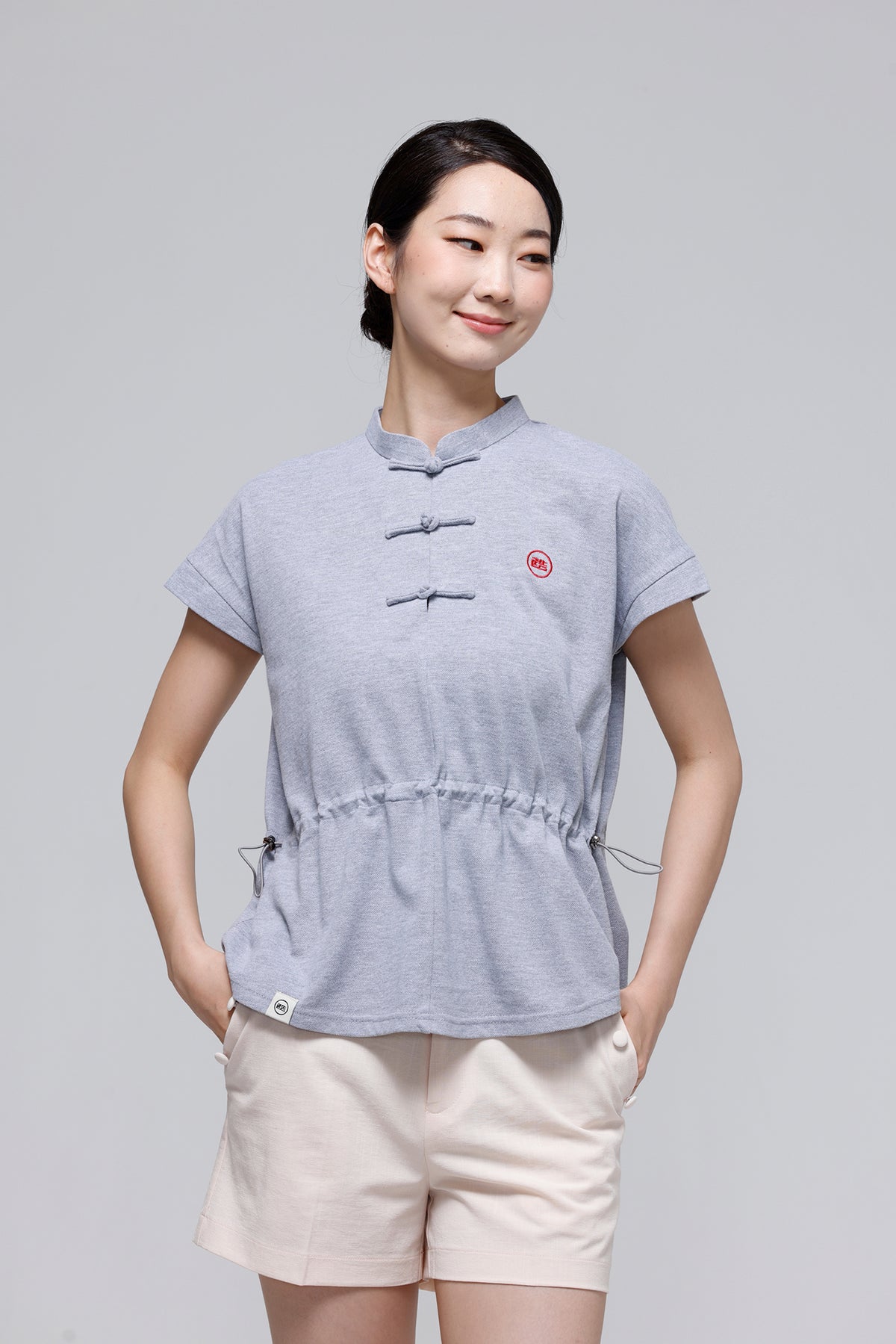 A woman modeling a light gray short-sleeve top with a traditional Chinese collar and three front toggle closures, paired with light beige shorts, against a solid gray studio background.