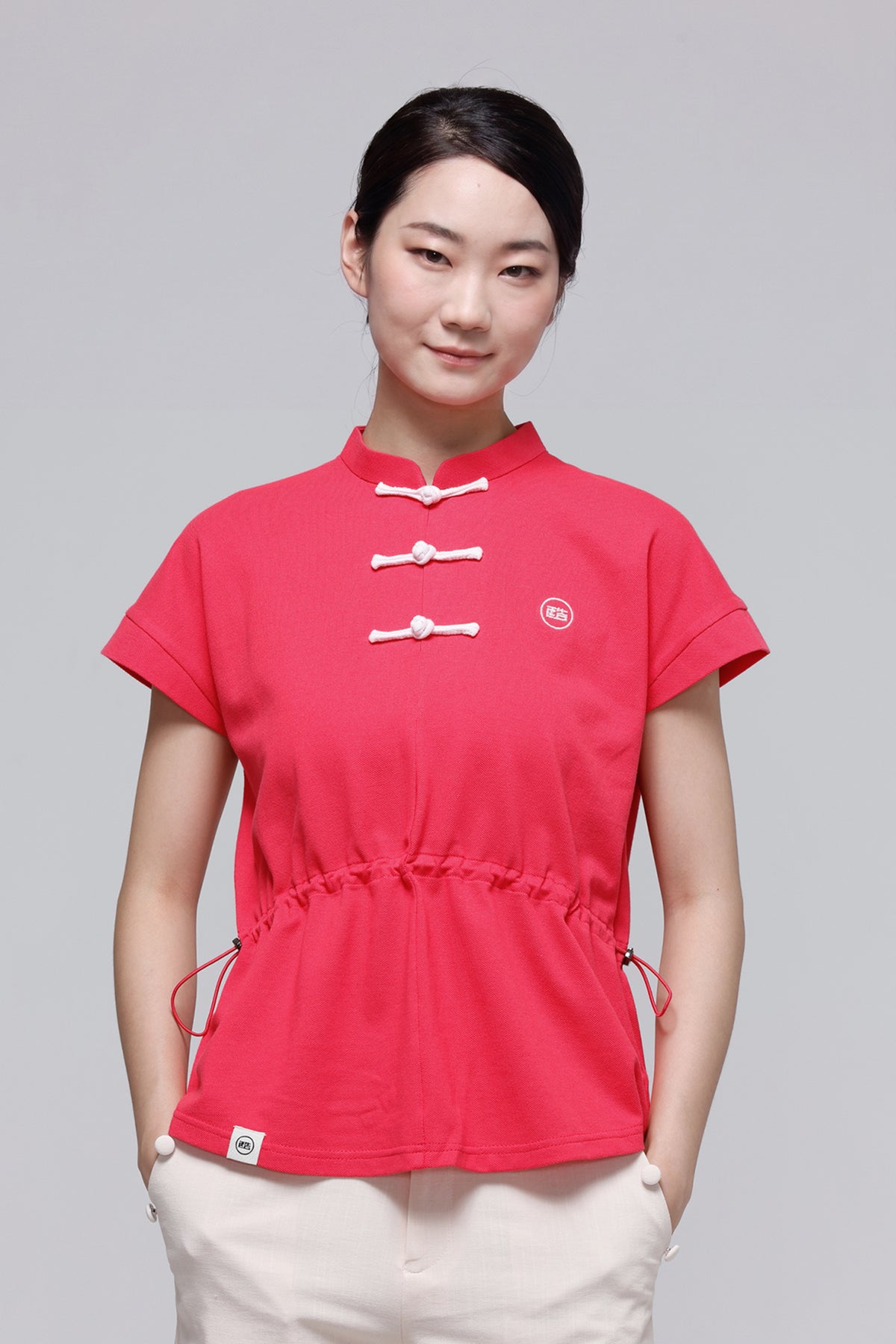 A woman modeling a bright pink short-sleeve top with a mandarin collar, three white Chinese-style toggle buttons, and a drawstring waist, paired with light beige pants against a solid gray background.