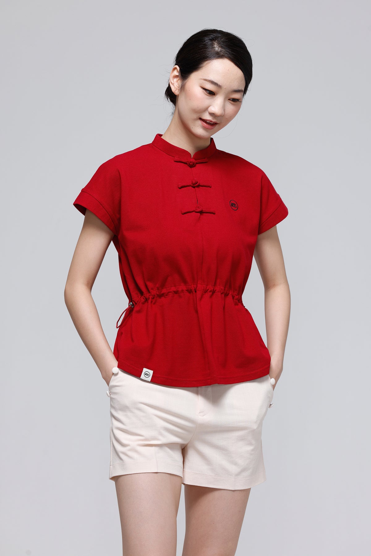 A woman modeling a red short-sleeve top with a traditional Chinese collar and black frog closures, paired with light beige shorts, against a solid gray studio background.