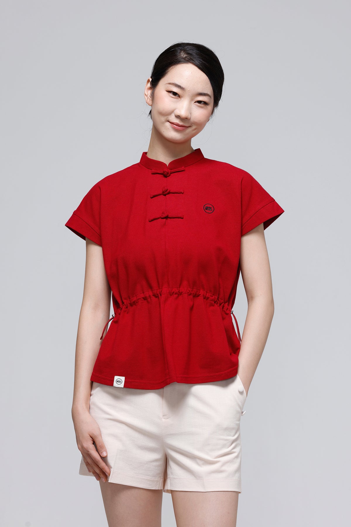 A woman modeling a red short-sleeve top with a traditional Chinese collar and black frog closures, paired with off-white shorts, against a solid light gray background.