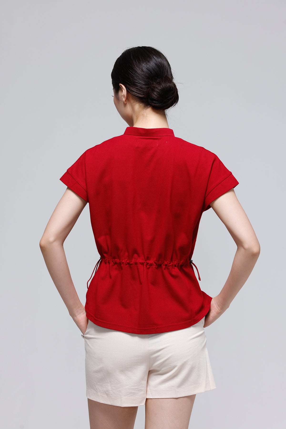 A woman viewed from behind wearing a red short-sleeve top with a gathered waist and drawstring, paired with light beige shorts, against a plain light gray studio background.