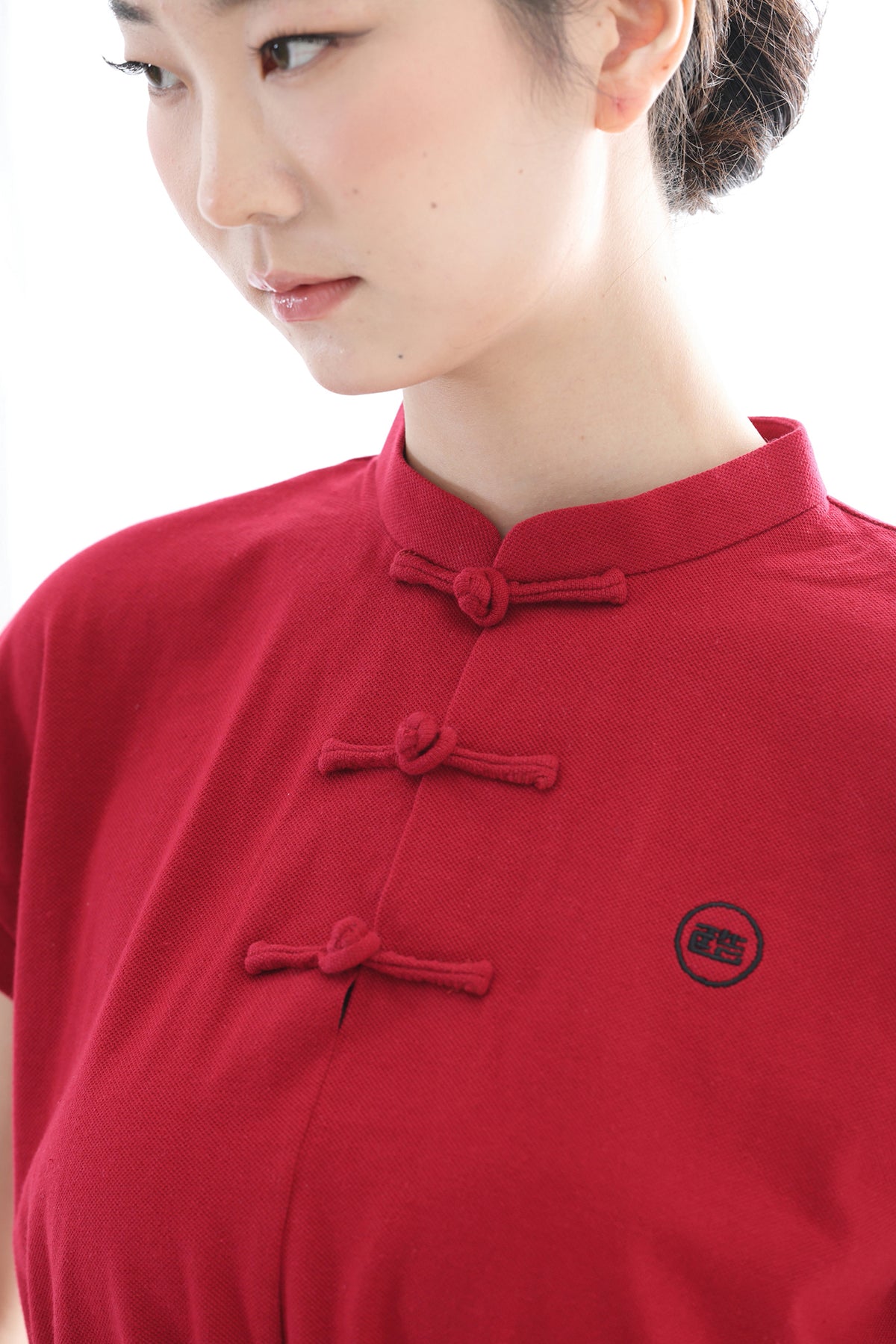 A close-up of a woman wearing a red short-sleeve mandarin collar top with traditional Chinese-style frog closures and a small circular black logo on the chest.