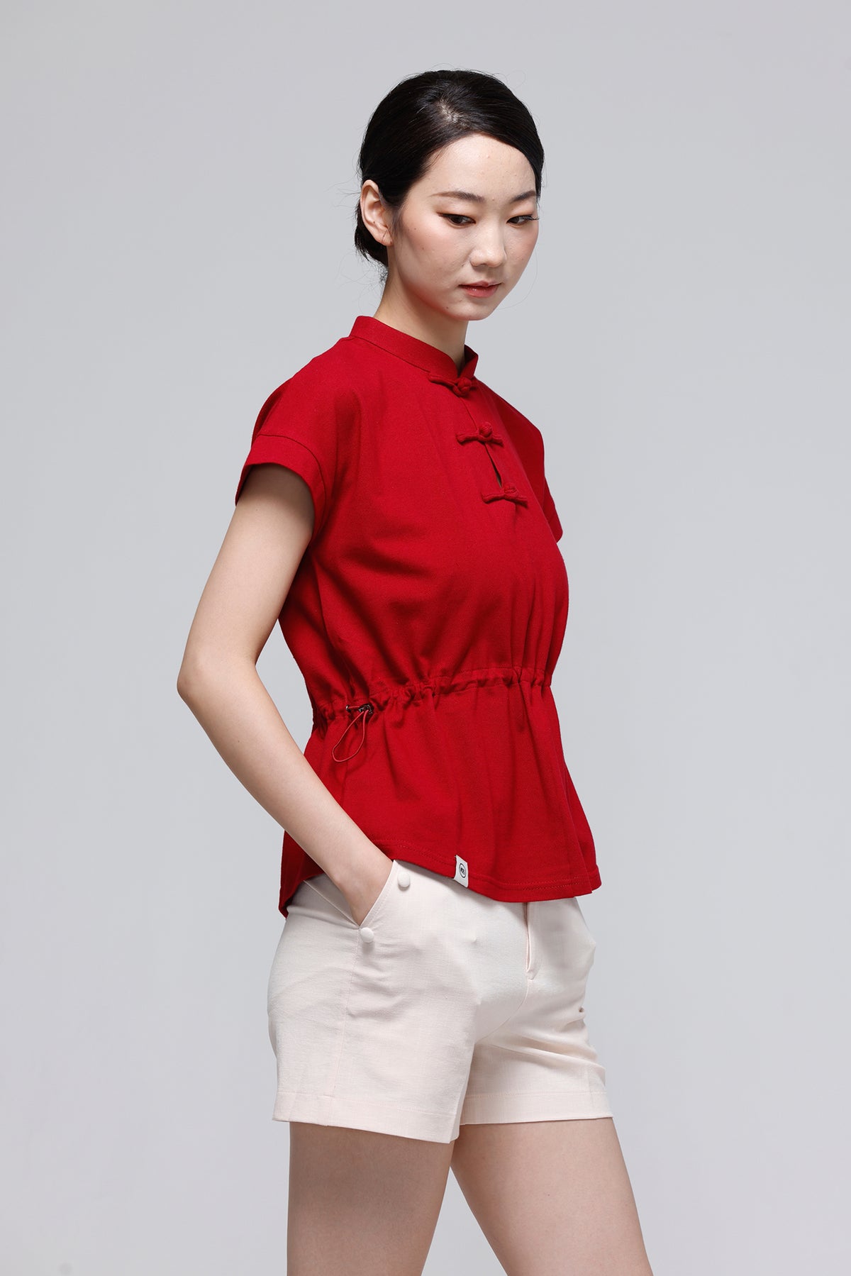 A woman modeling a red short-sleeve mock-neck top with black decorative embroidery and white shorts, standing against a light gray studio background.