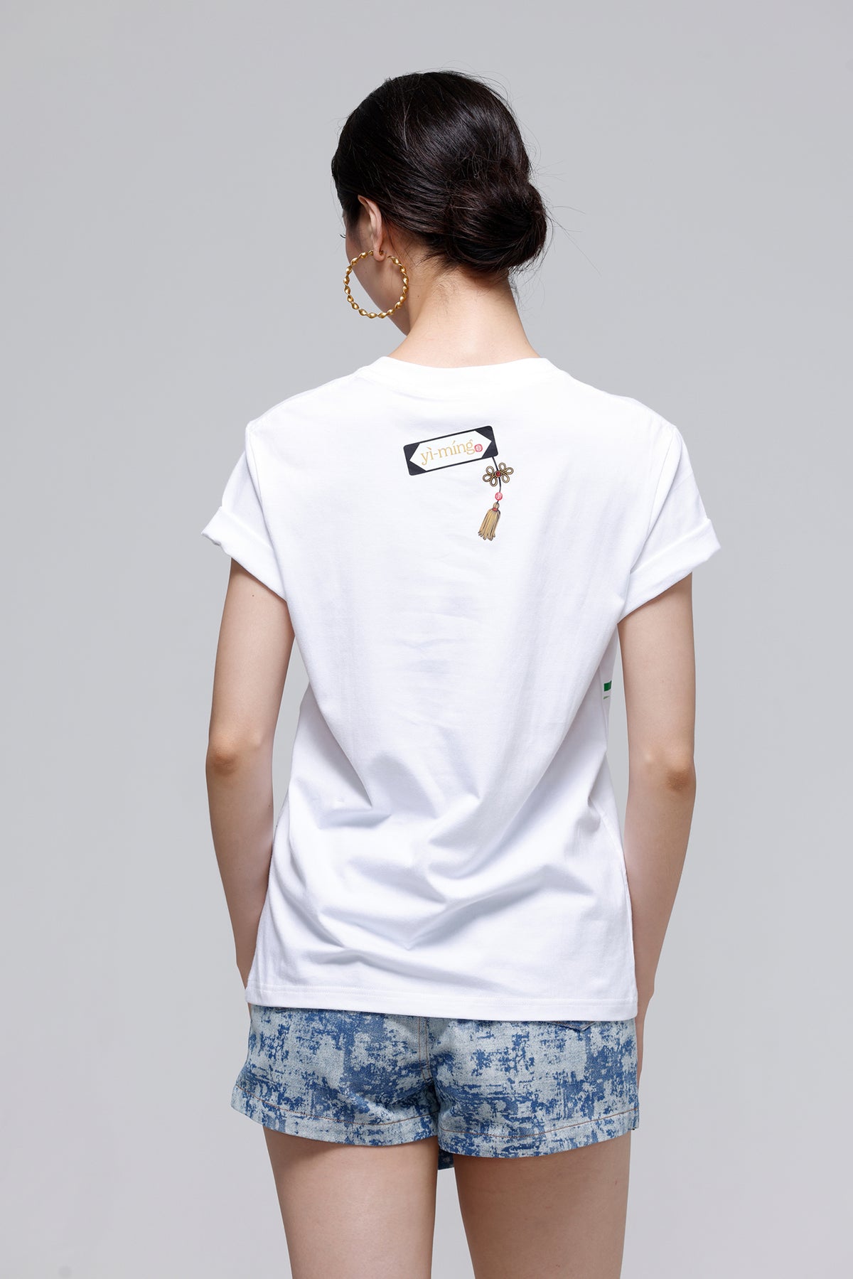 A woman viewed from behind wearing a white short-sleeve T-shirt with a small graphic print on the upper back and blue patterned shorts, against a plain light gray studio background.