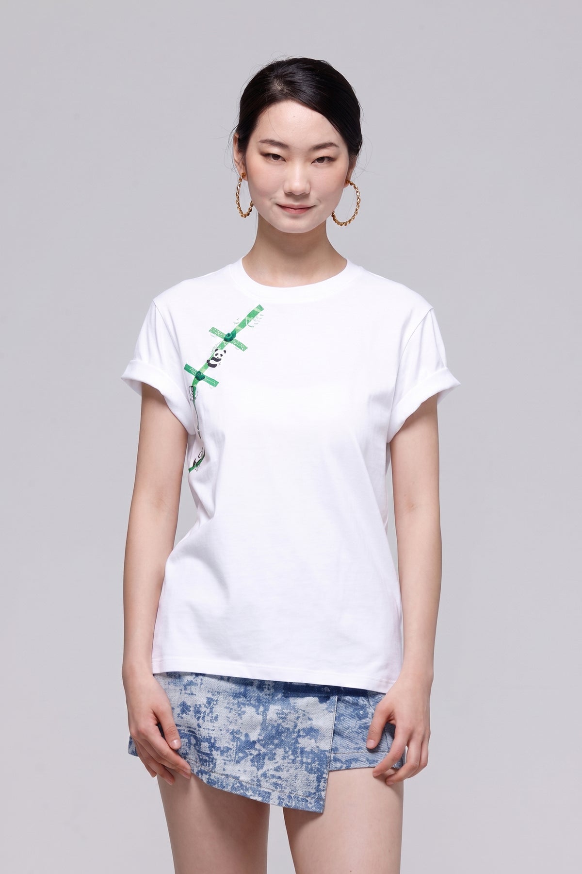 A woman modeling a white short-sleeve t-shirt with a green graphic print on the left chest, paired with a blue patterned skirt, against a solid gray studio background.