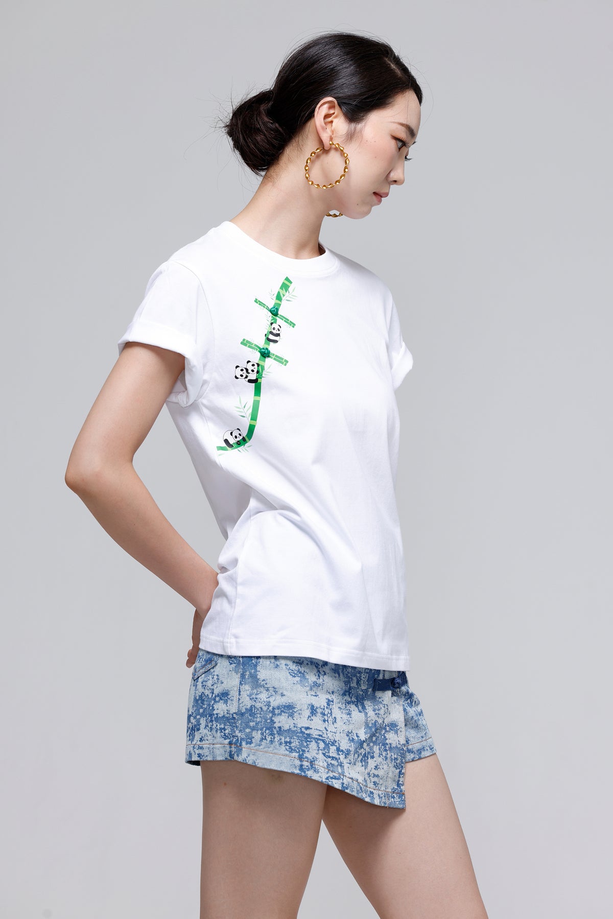 A woman modeling a white short-sleeve T-shirt with a green bamboo graphic and panda print on the front, paired with blue patterned denim shorts, against a solid gray studio background.