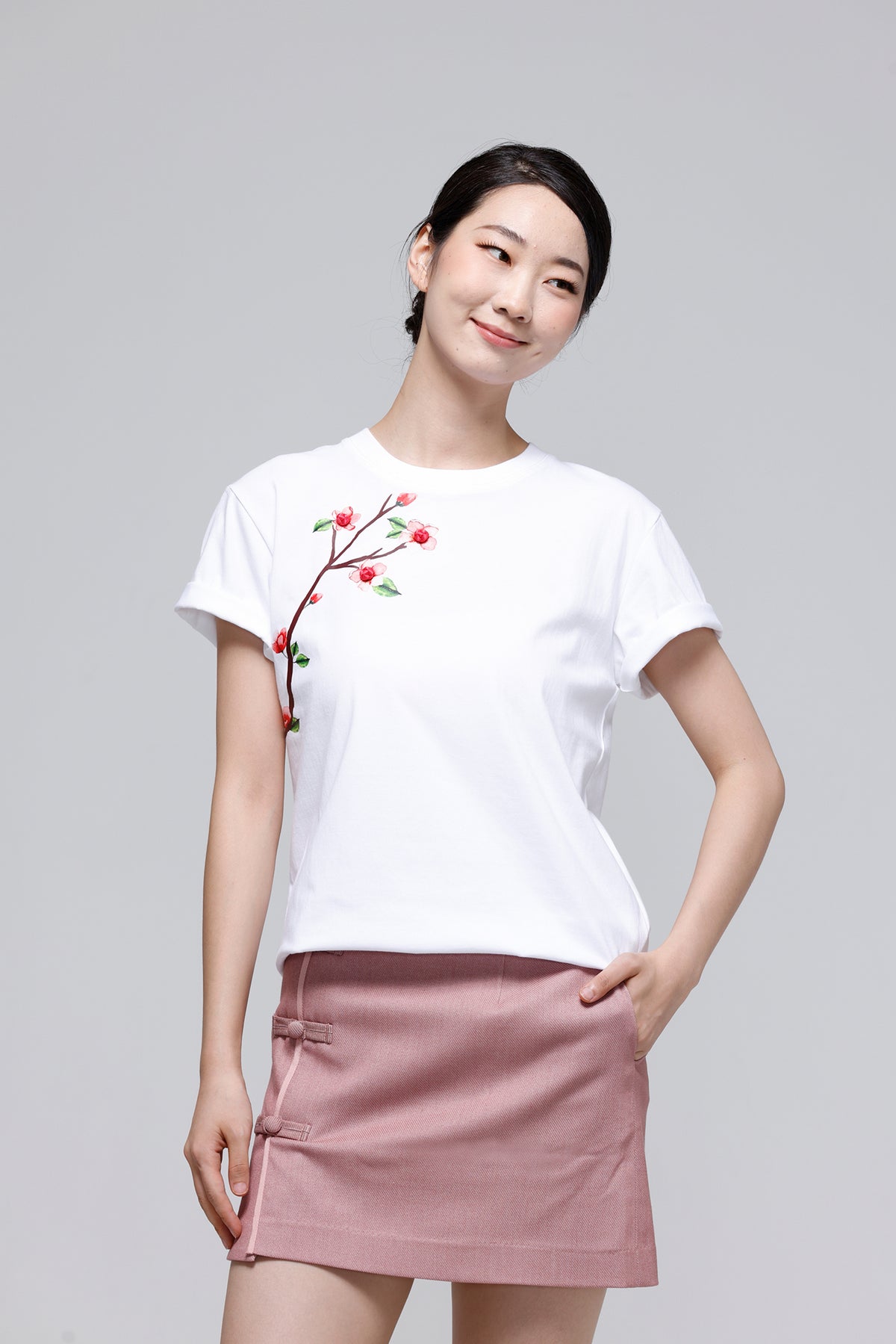 A woman modeling a white short-sleeve t-shirt with a red and green floral branch embroidery on the left chest, paired with a dusty pink mini skirt, against a solid light gray background.